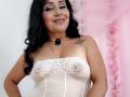 Zaria69 with black hair, smiling, wearing a sheer white lace top, showing her tits, black necklace, and star tattoo on her right shoulder. Pink background.