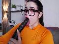 AryButty with dark hair and glasses, wearing an orange top, sucking a large black dildo with her tongue out, holding it with her right hand.