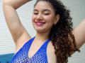 SanetPerlas with curly brown hair, arms raised, smiling, wearing a blue netted top, pink lipstick, small earrings, light skin, visible armpit, cute, confident, sexy.