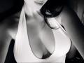 Olieri's tits are visible in a deep, white halter top with a plunging neckline, showing cleavage. Her face is partially out of frame, with dark hair. Black and white.