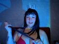 NaniSky with black hair, wearing a red bra, a silver crown, holding a black whip, tattoos on right arm, blue and white lighting, sitting on a red chair.