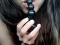 ImFireMoon's lips are around a black bead butt plug, holding it with her hand. Her brown hair is loose, and she has fair skin. Her tits are partially visible.