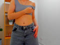 Steisii is wearing a gray one-shoulder top, lifting it to show her bare midriff, with blue denim pants, and has her hand on her crotch.