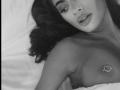Black-and-white photo of DariaAmaris lying down, topless, with a heart-shaped nipple piercing on her left breast. She has pouty lips and dark, wavy hair.