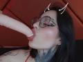 PaisleyMour is sucking a large, pale, circumcised dildo with pink lips, wearing glasses, and has dark tattoos on her neck. She has black hair and devil horns on her head.