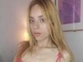 LyannaDifran has blonde hair, pink eyeliner, and pink lace bra showing her small tits. She's staring at the camera with a neutral expression. Soft lamp light in the background.