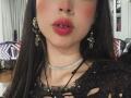 IsisNovafg with glossy red lips, a nose ring, and a jeweled choker, wearing black, torn lace top, and ornate earrings. Her dark hair is straight and long.