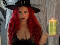 Estefy69 with red curly hair, black witch hat, and deep cleavage, holding a lit green candle, wearing black lace. Boobs, dark lipstick, and face paint dots.