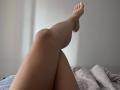 Lelliana's legs crossed at the ankles, with one leg raised, showing her bare feet and smooth, light skin. She's lying on a bed with a gray, patterned blanket.