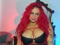 Estefy69 with red curly hair, glasses, and large black bra showcasing her tits, red strappy top, tattoo on right arm, smiling in a dimly lit room.