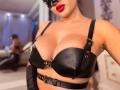 MademoiselleBella with big tits in a black leather bra, straps with gold buckles, red lipstick, black mask, and black gloves, in a blurred bedroom with a man in background.