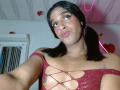 IdiiaLatina has dark skin and black hair in pigtails, looking into the camera with puckered lips. She wears a red, off-shoulder, mesh top with large, diamond-shaped cutouts revealing her chest. Her makeup includes small glitter dots on her cheeks. The background includes a white door, a pink neon heart, and a small red heart. The ceiling is white with visible panels. The image is a selfie with a slightly low angle. IdiiaLatina's top is shiny and textured.