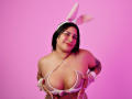 BonnieGray with white bunny ears, holding her large tits in a tiny white bikini top, smiling, pink background, black hair, tattoos on arms, white wristbands.