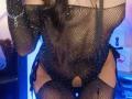 NahomyCarther wearing a black, sparkly fishnet bodysuit, exposing her pussy through a cutout, with long dark hair, a chain bracelet, and a sexy, confident pose.