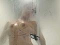 LilithDesire is standing in a shower, water spraying, hands covering her tits, visible through a fogged-up glass door. Her nipples are hard. She's naked, with water droplets.