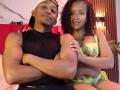 LilithAndAsmodeo, a muscular black man with a white cap, sits with arms crossed, wearing a black sleeveless shirt. Beside him, LilithAndAsmodeo, a curly-haired black woman, wears neon green lace lingerie.