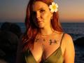 Rubbi has red hair, a white plumeria in her hair, green dress, tattoos on her chest, and a pensive expression, with her tits slightly visible, in sunset light.