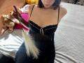Tisia with pale skin, wearing a black sheer dress with tiny white buttons, holding a white feather flogger, sitting on a bed with yellow and pink pillows.