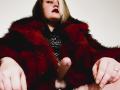 BeccaVanBoa in a red and black fur coat with a flirty lace top, exposing a big, erect, uncircumcised cock. She's looking up with dark lipstick and blonde hair.