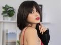 JasmineDay with dark brown hair, red lipstick, and yellow nails, wearing a red bra, black sleeve, touching her chin, looking at the camera, showing her tits.