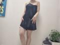 JoannaSia in a black, one-shoulder mini dress, hands on her hips, standing with legs apart, showing her bare ass and small tits, pale skin, and a potted plant.