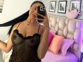 CassieFox with long black hair, wearing a sheer black lingerie dress, holding her hair, showing her tits, taking a selfie in a bedroom with pink pillows and a tufted headboard.