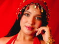 LilyConorr with shiny green eyes, glossy pink lips, and dark hair, wearing a red headpiece with gold coins, touching her cheek, showing off long, white nails. Red background.
