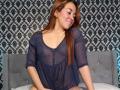 MadisonKoper with brown hair, smiling, wearing a sheer blue top, showing her small tits and nipples, sitting on a gray bed with a tufted headboard.