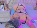 SashaPolet is on a bed, butt up, wearing black and white strappy lingerie, with blonde hair, a dildo in her butt, and a purple-blue light on her skin.