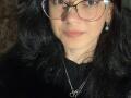 Dalaila with black wavy hair, wearing glasses, a black shirt, and a necklace, has fair skin and pink lips, looking straight at the camera with a slight smile.