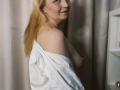 KarenVillis with blonde hair, white shirt off-shoulder, exposing small bare tits, side profile, smiling, light skin, curtain background, standing, right side, visible nipple, soft lighting.