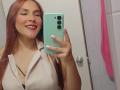 AntoniaBrunni with long red hair, smiling, holding a green phone, wearing a beige shirt, showing her tits, pink towel in the background, white tiled wall, door with knob.