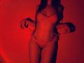 Tisia with long dark hair, wearing red lace bra and thong, pulling side strings, showcasing slim waist, small breasts, and flat stomach, under red lighting.