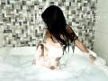 EstrellaXHot, nude, with wet black hair, sitting in a bubbly white bathtub, back to the camera, showing her ass and small tits, against a mosaic-tiled wall.