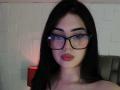 IsabellaaRossi with black hair, glasses, and red lipstick, has a nose ring, and a slightly serious expression. Her tits are not visible. She's against a white textured wall.