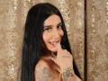 SamiSins with long black hair, smiling, showing teeth with finger near mouth, tattoos on arm, gold bracelet, brown eyes, cute, boobs visible, glossy curtain background.