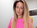 ViktoriiaLu hat langes, blondes Haar, trägt einen hellrosa Pullover und wirkt leicht verärgert. Sie hat einen kleinen Nasenring in ihrem linken Nasenloch und befindet sich in einem schlichten Innenraum..