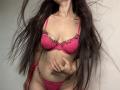 Tisia with long brown hair, pink lace bra, and matching pink lace panties, showing her tits and pussy, hand raised, standing against a plain wall.