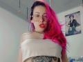 NatashaLau with bright pink and black hair, glasses, red lipstick, off-shoulder white top, tattoos, necklace, and red earrings, looking at camera with pouty lips.