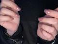 MorganCastellanozs Hände sind mit schwarzen Metallfesseln gefesselt, wobei glänzende, nackt lackierte Nägel mit einem dezenten Rosaton zu sehen sind. Die Hände sind nach vorne positioniert, die Finger leicht gekrümmt..