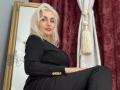 Hotsexydolly with platinum blonde hair, red lipstick, black dress with gold buttons, sitting in front of a mirror, white curtain with gold tassel, maroon velvet backdrop.