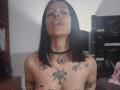 AmelieBlake with black hair, nose ring, and purple gem necklace, topless, tongue out, showing tattoos on chest and arms, small tits, and a large bat tattoo on chest.