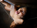 CatalinaRivero with wet black hair, tongue out, holding a showerhead, water on her tits, brown eyes, and earrings, kneeling on a dark floor, looking up.