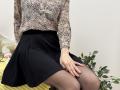 Rorria wearing a textured, button-up blouse, black skirt, and sheer black pantyhose, sitting with her hand on her thigh, showing her panties' outline.