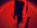 Tisia's back is to the camera, arms raised, long dark hair flowing, wearing a red bra, her ass slightly visible, bathed in red light, creating a sensual, sexy vibe.