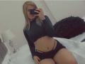 MalloryHearth with blonde hair, black crop top, and black shorts, holding phone, showing her flat stomach and small ass, sitting on bed, black pillow in background.