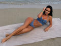 SexyGirl with tan skin, black hair, and small tits, lying on a towel in blue plaid bikini, smiling, hand in hair, beach, legs stretched out, water in background.