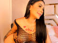 CristalRousse with long black hair, smiling, wearing a black fishnet top, exposing her tits, tattoo of a heart and "obuJ" on her chest, and tattoos on her arm.