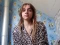 BessBetak with long, dirty blonde hair, wearing a leopard print shirt, looking straight ahead with a neutral expression, in a room with blue, cartoon-patterned wallpaper.