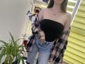 Nesie with bare tits in a black strapless top, denim shorts, and a pink plaid shirt off her shoulders, standing by a green plant and yellow blinds.